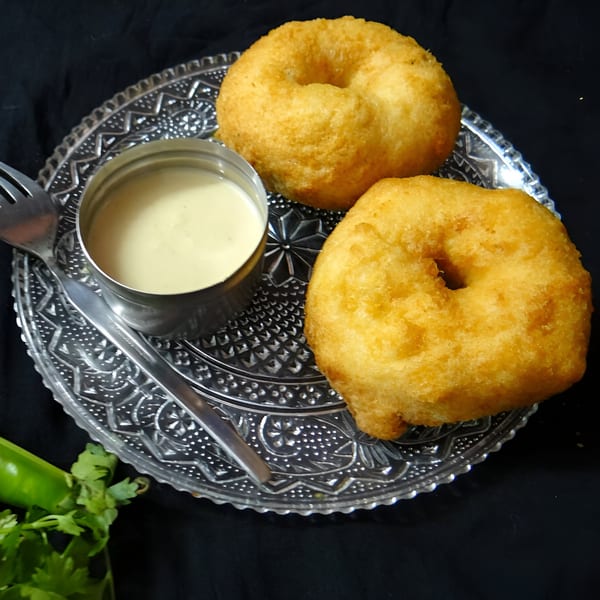 Two golden-fried medu vada, a small bowl of chutney, and a fork on a clear glass plate.