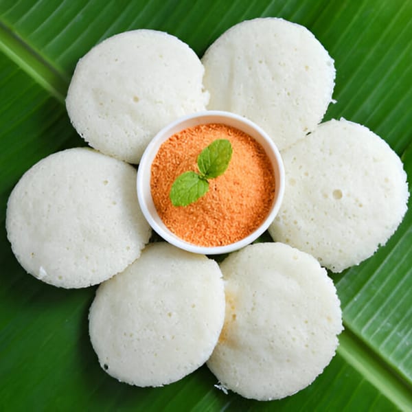 Six white idlis arranged around a bowl of orange chutney with mint on a banana leaf.