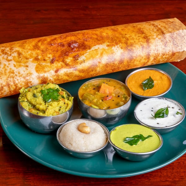 A golden-brown dosa roll with six small bowls of colorful Indian chutneys and sambar on a teal plate.